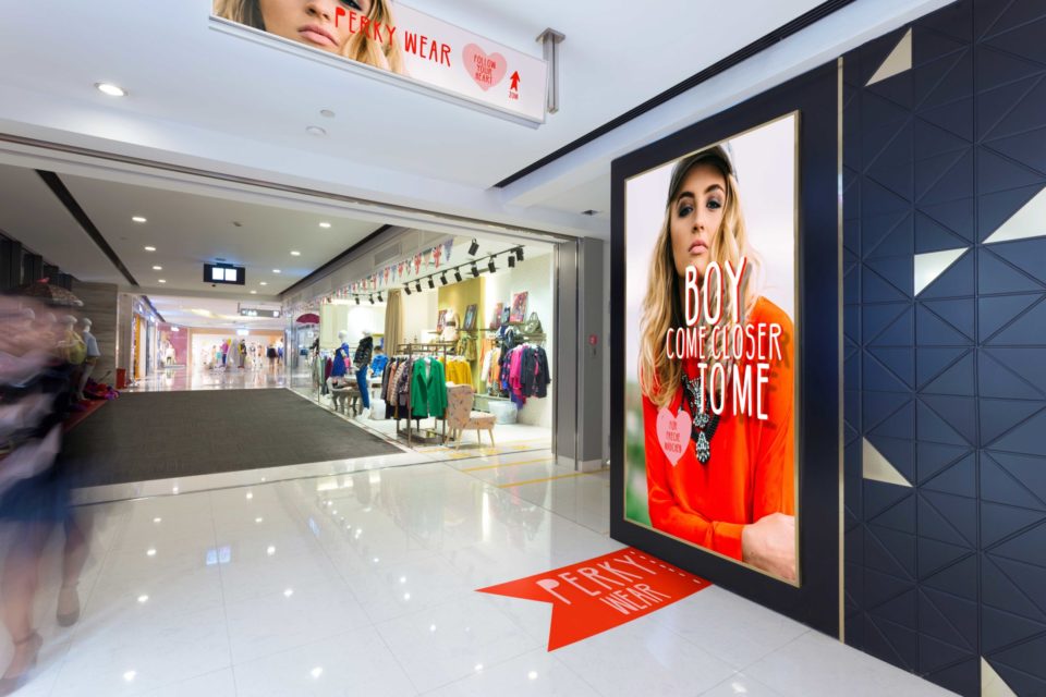 Backlit advertisement with woman dressed in red in shopping arcade next to shop entrance