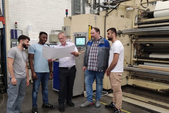 2 trainers look at paper in front of coating machine with 3 male apprentices